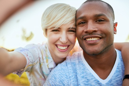Mixed race couple of millennial in a grass field taking a selfie with a smartphoneの写真素材