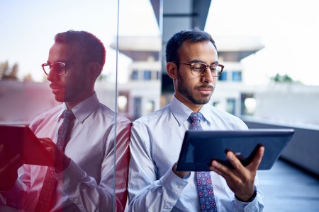 Millenial businessman leaning confidently on a dark glass wall with cityscape backgroundの写真素材
