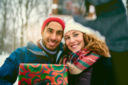 Smiling diverse couple holding Christmas presents and holding a mobile phone to take a selfie while...