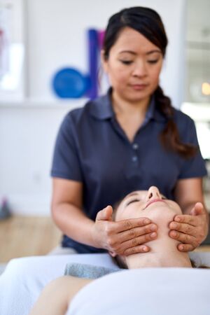 Chinese woman massage therapist giving a treatment to an attractive blond client on massage table in a bright medical officeの写真素材