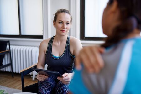 Caucasian woman physiotherapist talking to a mid-adult chinese female patient and taking notes on a tablet during a fitness evaluation consultationの写真素材