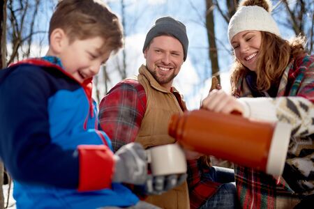 Family with son enjoying playing in fresh snow during wintertime and having hot chocolate during a picnicの写真素材