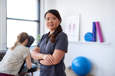 portrait of a Chinese woman massage therapist giving a neck and back pressure treatment to an attractive blond client at her workplace in a bright officeの写真素材