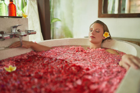 Gorgeous millennial girl pampering herself in modern bath tub filled with flower petals in tropical...