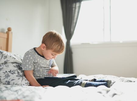 Adorable male kid wearing trendy tshirt sitting on bed using digital tablet with young parent in modern home with pet dog, practicing reading and writingの写真素材