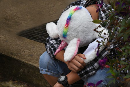 A young man in a black-and-white plaid shirt and blue shorts, in a white baby cap in the shape of a unicorn's head, sits on a step in the street, his head down, his face is not visible.の写真素材