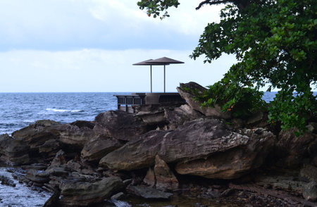 View from the rocky coast to the sea, in the foreground large stones and braided sofas under two beach umbrellasの写真素材