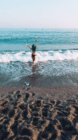 Happy young white girl running hands up into the sea. Carefree stress free lifestyle. Finally vacation near the ocean. Joyful adult woman excited to swim on a relaxed evening on the beach at sunset.の写真素材