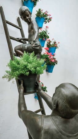 Cordoba / Spain - October 2019: Famous sculpture: a boy climbing a ladder and a man giving him a flower pot to decorate a white wall outside of a house. Close up view. Spring patio decorating festivalのeditorial素材