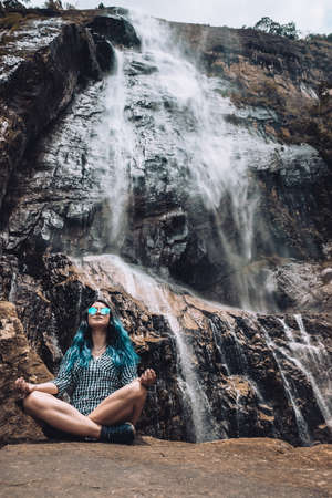 Young white girl sitting under Diyaluma waterfall, Sri Lanka. Happy woman in meditation pose on vacation. Wellness lifestyle, healthy yoga retreat in summer. Peaceful holiday in beautiful exotic placeの写真素材