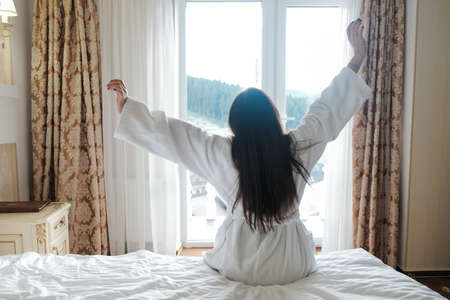 Young brunette girl stretching in hotel room bed in the morning. Waking up full of energy on vacation, arms raised and fully awake. Happy and lazy early day. Female sitting in bathrobe on a retreat.の写真素材