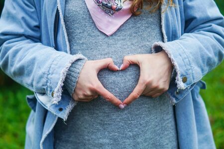 pregnant woman in jeans jacket holding her hands in a heart shapeの写真素材