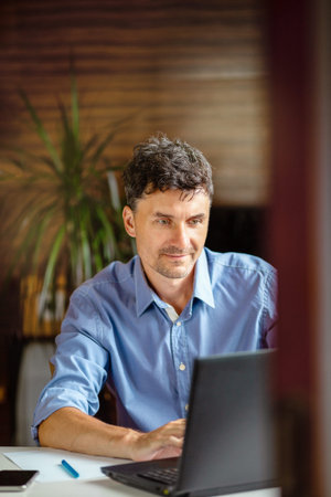 Middle-aged man runs a business from home. Works at a computer in a room with a home environment....
