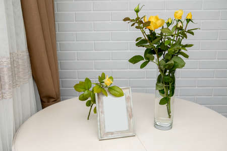 Blank photo frame and yellow roses in the vase standing over the white wall. Place for your textの写真素材
