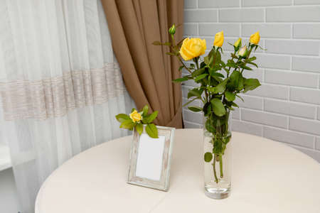 Blank photo frame and yellow roses in the vase standing over the white wall. Place for your textの写真素材