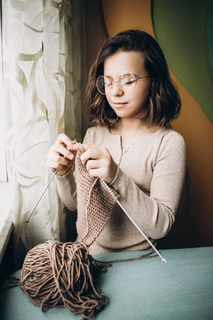 Teenage girl in a beige sweater, wearing glasses knitting needles standing by the window. The concept of hobbies, creativity, needlework, handwork.の写真素材