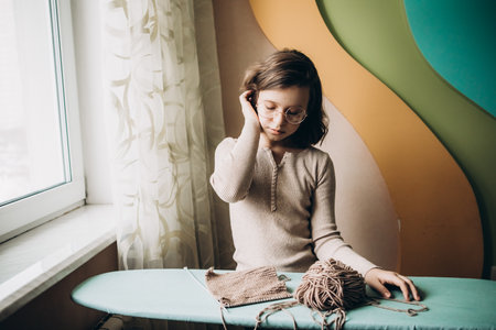 Teenage girl in a beige sweater, wearing glasses knitting needles standing by the window. The concept of hobbies, creativity, needlework, handwork.の写真素材