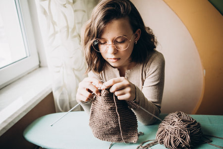 Teenage girl in a beige sweater, wearing glasses knitting needles standing by the window. The concept of hobbies, creativity, needlework, handwork.の写真素材