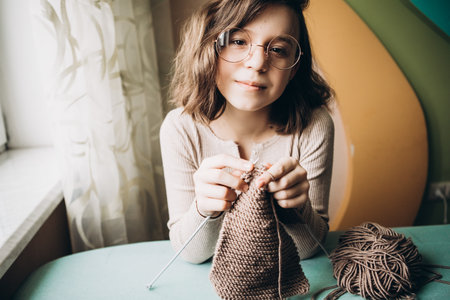 Teenage girl in a beige sweater, wearing glasses knitting needles standing by the window. The concept of hobbies, creativity, needlework, handwork.の写真素材