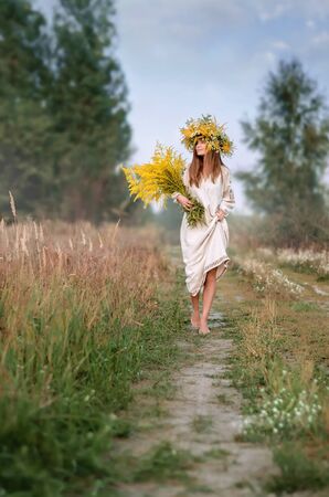 Beautiful girl in a wreath of wildflowers and a ancient embroidered clothes with a large bouquet walking barefoot on the path. Ukrainian culture, holiday of Ivana Kupalaの写真素材