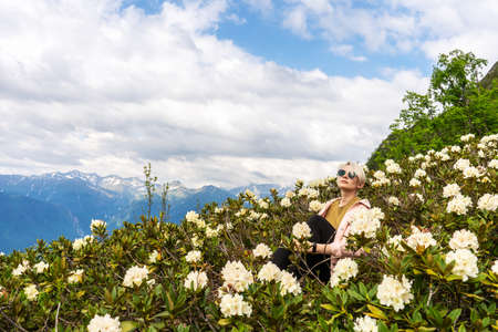 A wonderful summer day in the mountains. The green slopes of the mountains are strewn with flowers. A young woman is sitting in flowers.の写真素材