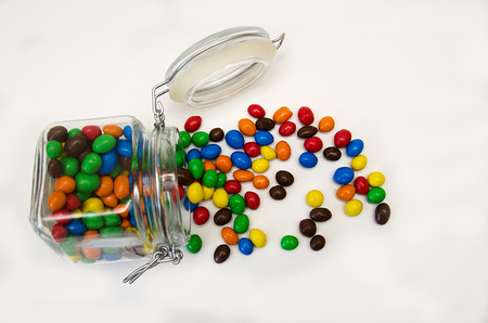 colored sweets, in red, orange, yellow, green, brown, blue glaze, lie scattered from transparent jar with lid on white backgroundの写真素材