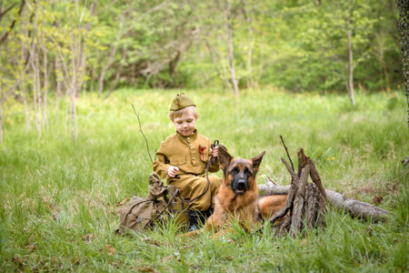 a boy in a military uniform in a clearing, sitting by a campfire with a German shepherd.Two friends defend the motherlandの写真素材