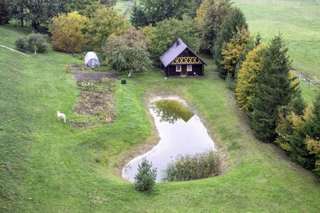 Wooden house in nature in the field. Country house with cow grazing in front and rectangular pond. Top view. の写真素材