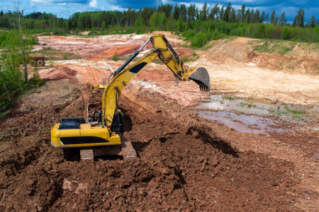 In a quarry with red clay, a yellow excavator with a large bucket.