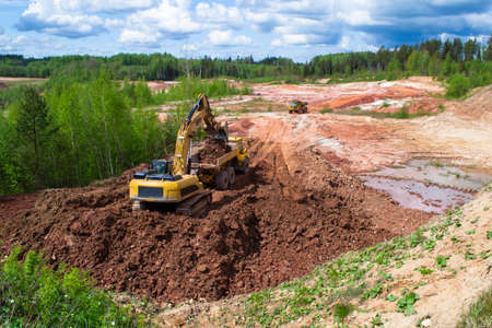 Clay quarry. The yellow excavator digs red clay with a large bucket and puts it into the truck.の写真素材