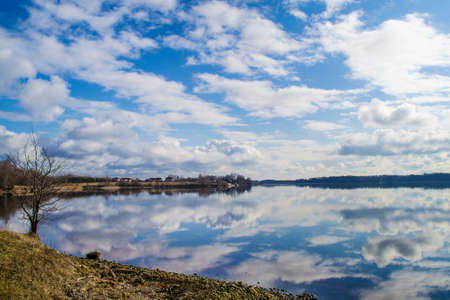 Tree and rocky shore in the foreground. Picturesque view of the Daugava. The nature of Latvia. Beautiful clouds in the blue sky and reflected in the waterの写真素材