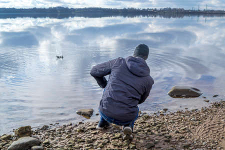 A man sits on the bank of a river and throws stones into the water. Dressed in a warm jacket and hat. Beautiful clouds in the blue sky and reflected in the water.の写真素材
