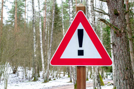 Winter road in forest on background. Road sign triangular shape with exclamation mark. Red with white attention traffic sign.の写真素材