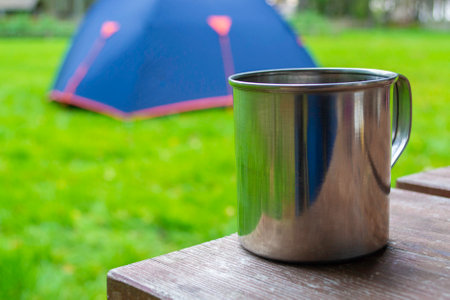 Close-up of travel cup. Metal mug with tea or coffee on wood table. Blue touristic tent at background. Copy space.の写真素材