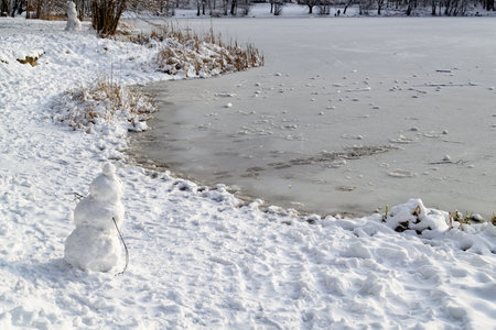 A small snowman stands on the shore of the lake. Snowy forest and frozen lake. Concept of winter games and New Year's mood.の写真素材