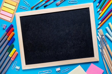 School supplies make frame around black chalkboard. Blackboard in the center. Colorful pencils, felt-tip pens, brushes, stickers and stationery flat lay on blue background. .Back to school conceptの写真素材