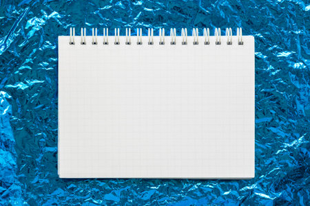 Notebook with spiral lie on background of blue crumpled foil. Blank empty sheet of white paper with...