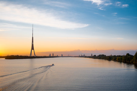 Riga old town panorama and TV tower. Colorful sunset over the Daugava river. A motor boat floats on...