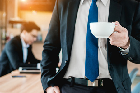 Businessman holding a white coffee cup and drink coffee in the morning. Another businessman working behind him.の写真素材