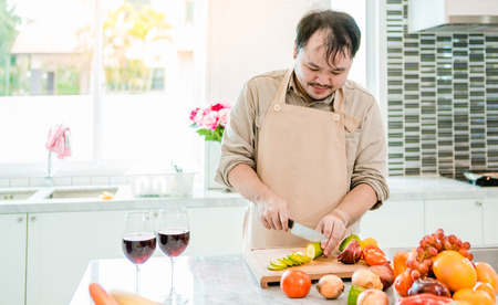 The man is cutting the cucumber in the kitchen in his house to prepare salad along with eating dinner.の写真素材