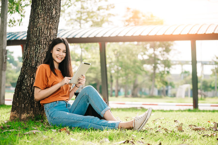 A young or teen asian girl student in university smiling and reading the book and look at the tablet or labtop computer in summer holiday.の写真素材