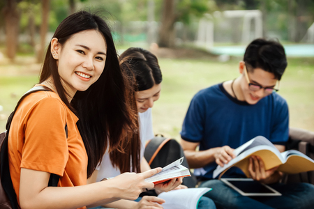 A group of young or teen Asian student in university smiling and reading the book and look at the tablet or laptop computer in summer holiday.の写真素材