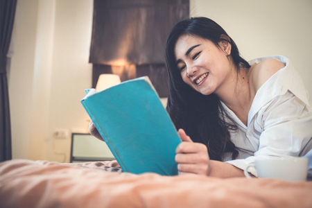Asian girl who just wake up in the morning as relaxed. She's read one book on the bed. She opened the window to receive the light of the morning sun. She was holding a glass of coffee to drink it.の写真素材