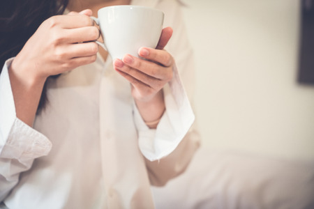 Asian girl who just wake up in the morning as relaxed. She's read one book on the bed. She opened the window to receive the light of the morning sun. She was holding a glass of coffee to drink it.の写真素材