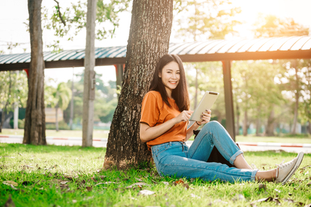A young or teen asian girl student in university smiling and reading the book and look at the tablet or labtop computer in summer holiday.の写真素材