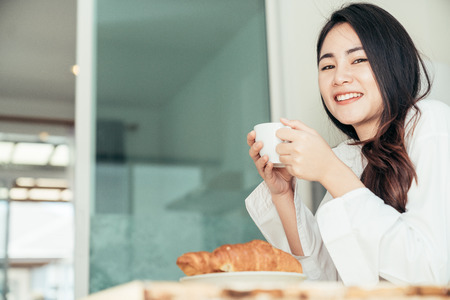 Asian girl who just wake up in the morning as relaxed. She was holding a cup of coffee or tea to drink it and eating the bread.の写真素材