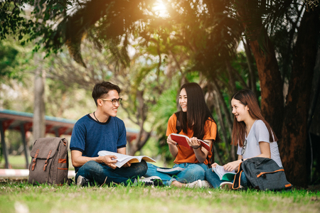 A group of young or teen Asian student in university smiling and reading the book and look at the tablet or laptop computer in summer holiday.の写真素材