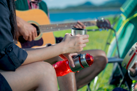 A cropped photo of  group of Asian friends sitting on chairs, singing and drinking some beer and water together outside the tent while they has camping on Weekend holiday.の写真素材