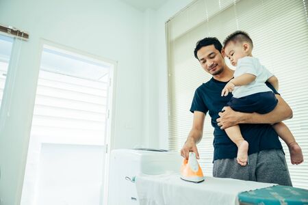Asian men are father ironing clothes and carrying his son or little boy in their home on holiday.の写真素材