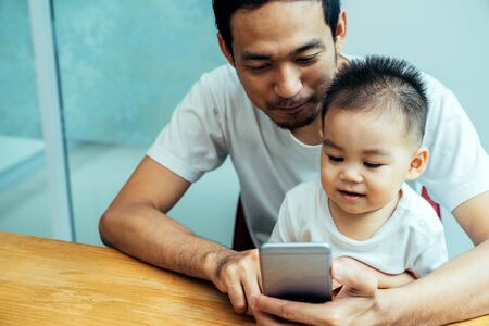 Asian father and his son is using the mobile phone at their home on sunday or holiday.の写真素材